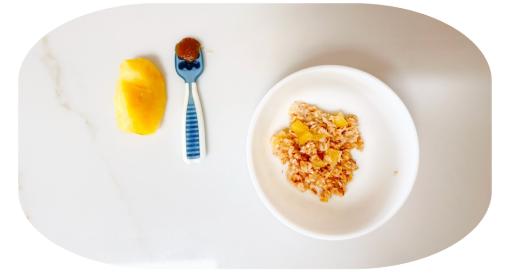 Little Bellies How to prepare apples for 6–7 month old babies | Little Bellies AUS Breakfast scene: bowl of granola with mango pieces on a white plate, plus a pineapple chunk and a spoon with jam on a white surface.