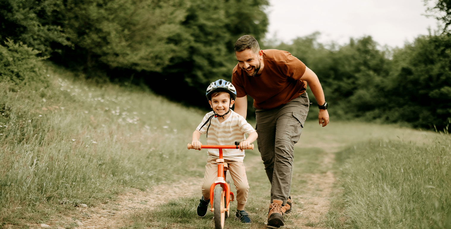A dad pushing his child, teaching him how to ride a bike in a field
