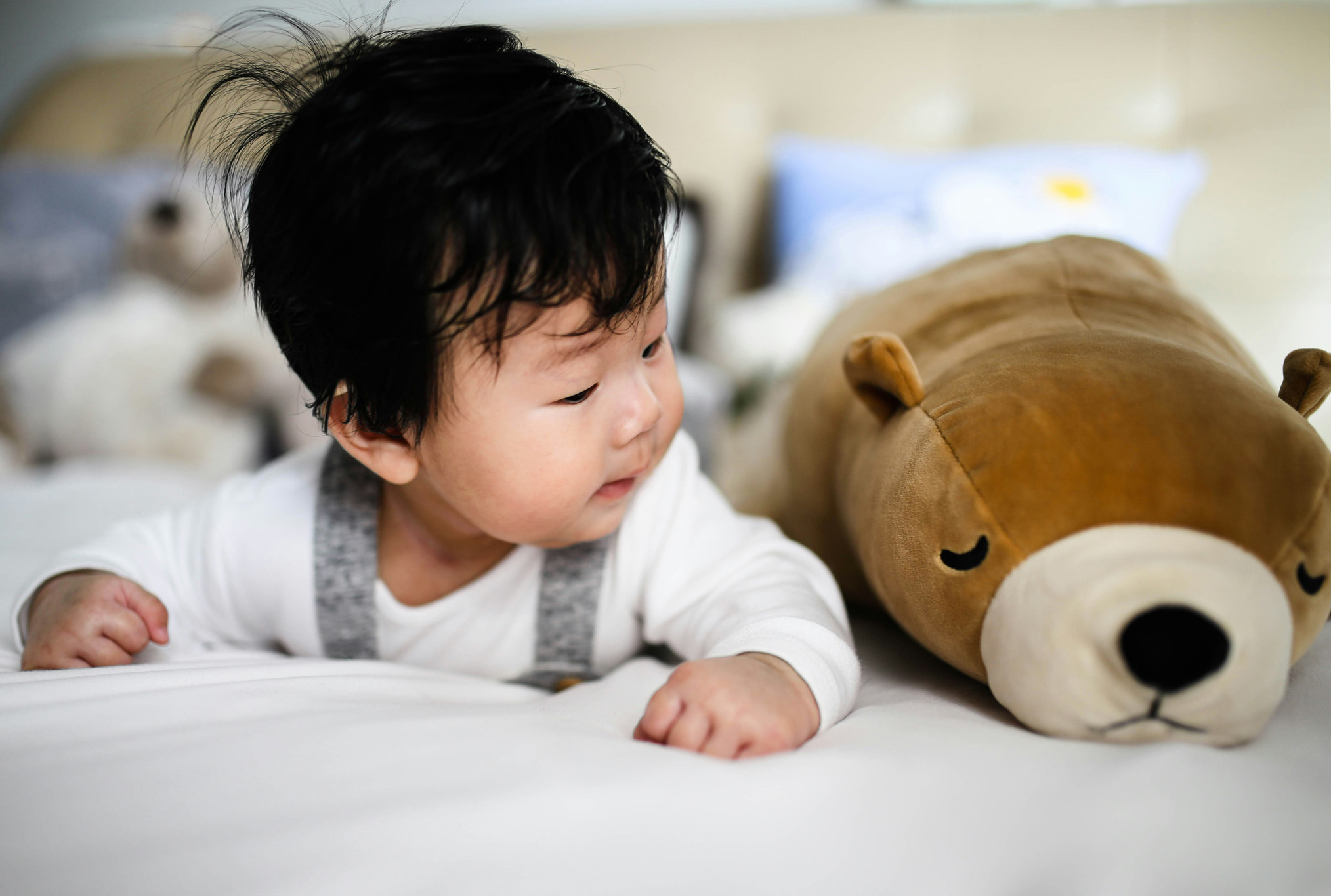 Young child lying on a bed beside a brown stuffed bear, looking at it curiously.