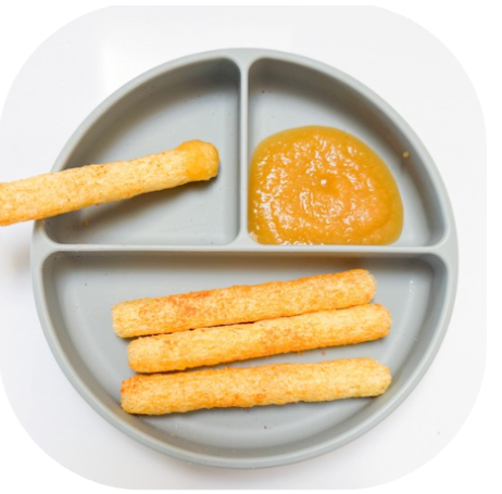 Apple purée with Little Bellies Organic Sweet Potato Pick-Up-Sticks | Little Bellies NZ Round divided plate with orange dipping sauce in the top-right compartment and four cheese sticks on the bottom; one cheese stick rests along the left edge.