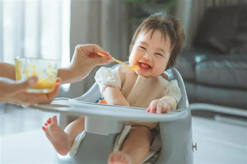 A baby in a high-chair laughing and being fed some food on a spoon. First Foods & Allergens: What to Know as You Begin